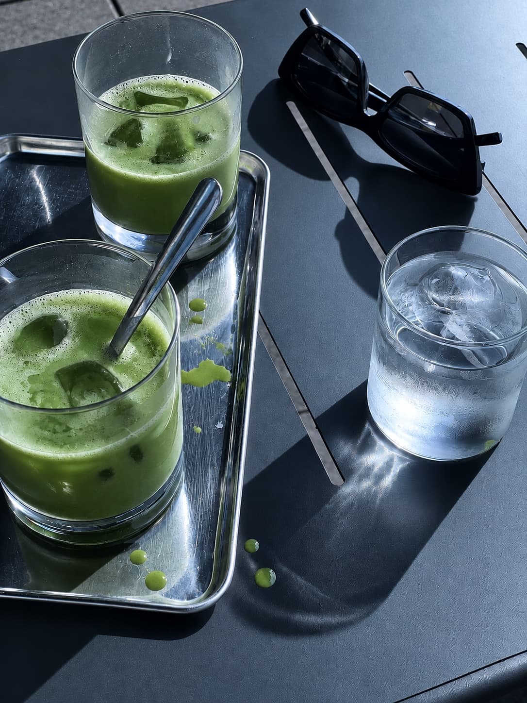 Dois copos de matcha gelado numa mesa escura, com óculos de sol e um copo de água